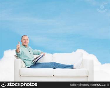 home, leisure and happiness concept - smiling man lying on sofa with book and showing thumbs up