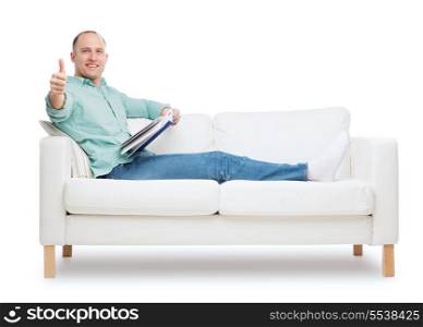 home, leisure and happiness concept - smiling man lying on sofa with book and showing thumbs up