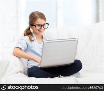 home, leisure, technology and internet concept - little student girl in eyeglasses with laptop computer at home