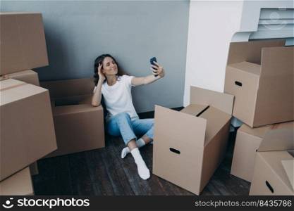 Home owner is taking selfie in new luxurious apartment. Happy woman unpacking cardboard boxes. Girl is taking photo on phone and sitting on the floor. Independence and success concept.. Happy woman unpacking boxes and taking selfie in new luxurious apartment. Success concept.
