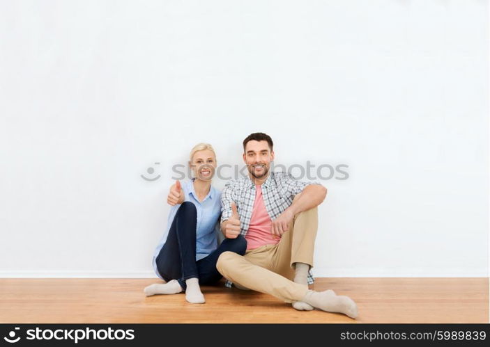 home, people, repair, moving and real estate concept - happy couple sitting on floor and showing thumbs up at new place