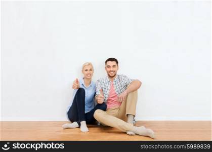 home, people, repair, moving and real estate concept - happy couple sitting on floor and showing thumbs up at new place