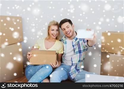 home, people, technology and real estate concept - smiling couple with cardboard boxes moving to new place and taking smartphone selfie over snow