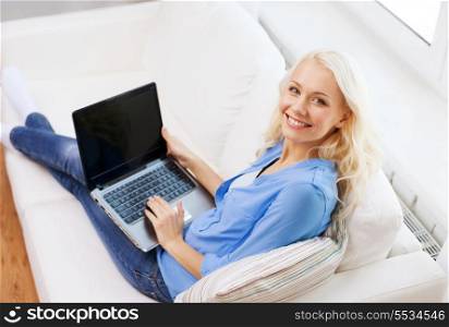 home, technology and internet concept - smiling woman sitting on the couch with laptop computer with blank black screen at home
