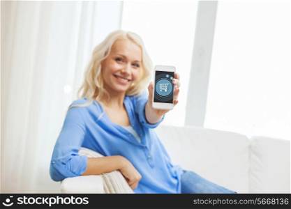 home, technology and people concept - smiling woman with showing smartphone screen with shopping trolley icon sitting on couch at home