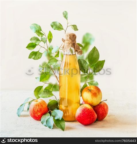 Homemade apple juice in glass bottle standing on white table with red apples and green leaves at white wall background. Healthy fermented food. Apple preserving