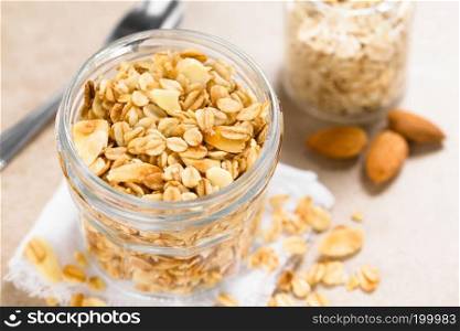 Homemade baked crunchy oatmeal, sliced almond, honey and coconut oil breakfast granola in glass jar, ingredients and spoon in the back  Selective Focus, Focus in the middle of the granola in the jar . Oatmeal, Almond and Honey Granola