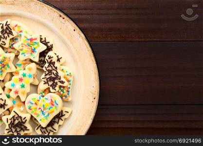 Homemade baked sugar cookies for Christmas with icing and colorful sprinkles on the top, served on enamel plate, photographed overhead on dark wood. Colorful Sprinkled Christmas Sugar Cookies