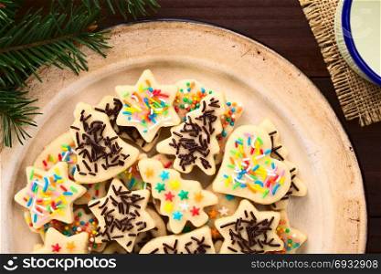 Homemade baked sugar cookies for Christmas with icing and colorful sprinkles on the top, served on enamel plate, cup of milk on the side, photographed overhead on dark wood. Colorful Sprinkled Christmas Sugar Cookies