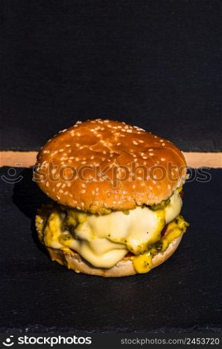 Homemade beef cheeseburger with melting cheese, tomatoes, onions, pickles isolated on a black stone plate.. Homemade beef cheeseburger with melting cheese, tomatoes, onions, pickles isolated on a black stone plate.