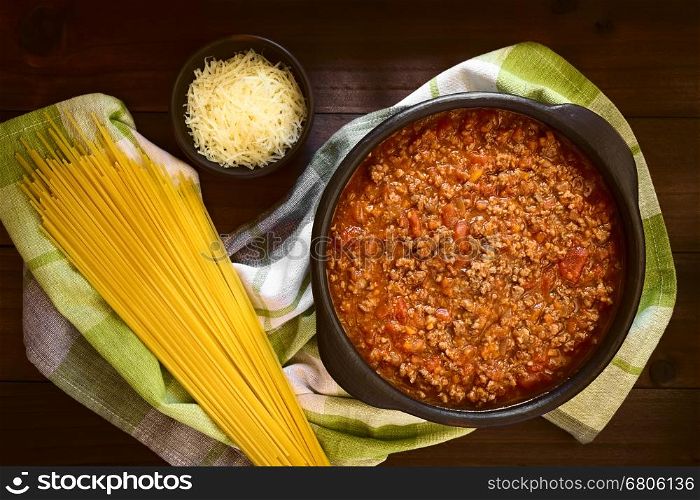 Homemade bolognese sauce made of fresh tomatoes, onion, carrot, garlic and mincemeat served in rustic bowl, uncooked spaghetti and grated cheese on the side, photographed overhead on dark wood with natural light