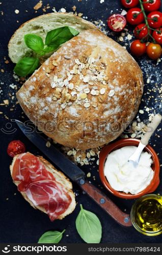 Homemade bread loaf and fresh ingredients for making sandwiches (tomatoes, basil, olive oil, cream cheese) on rustic dark background