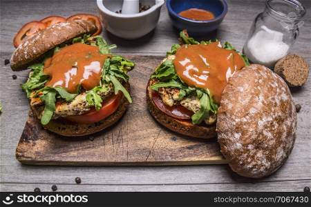 homemade burgers with chicken in mustard sauce, arugula and tomatoes on a cutting board with spices and sauce on wooden rustic background top view close up