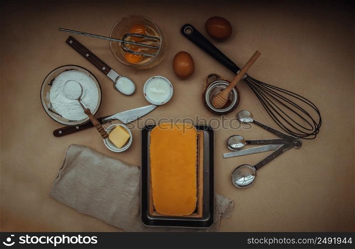 Homemade Butter cake or Pound cake with ingredients (eggs, flour, milk, butter with honey) and rolling pin, egg whisk on light brown table. Top view, Selective focus.
