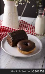 homemade chocolate donut and sponge cake covered with chocolate glaze and glass of milk on white rustic wooden surface