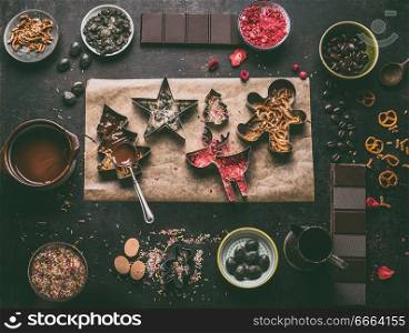 Homemade Christmas chocolate bars making. Christmas cutters with various toppings and flavorings. Melted chocolate in bowl with spoon on dark rustic kitchen table background, top view. Edible gifts