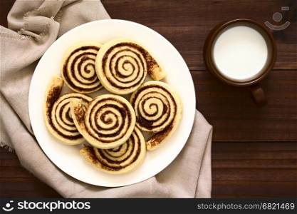 Homemade cocoa rolls or buns made of yeast dough and filled with sugar and cocoa powder, cup of milk on the side, photographed overhead on dark wood with natural light