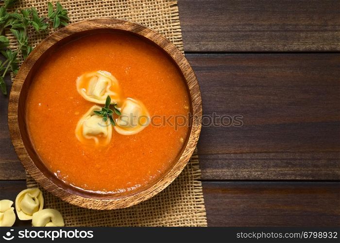 Homemade cream of tomato soup with tortellini garnished with fresh oregano, served in wooden bowl, photographed overhead on dark wood with natural light (Selective Focus, Focus on the top of the soup)