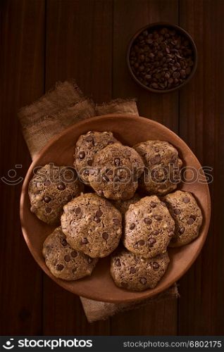 Homemade double chocolate chip cookies on rustic plate with chocolate chips in small bowl above, photographed overhead on dark wood with natural light