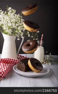 Homemade doughnuts covered with chocolate glaze falling on white plate and glass of milk on white rustic wooden surface. Levitation photography