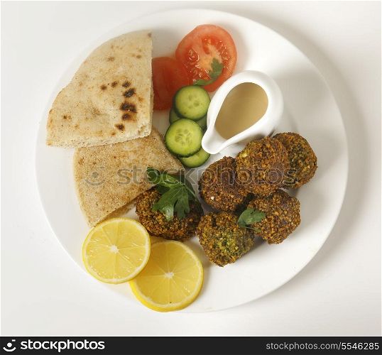 Homemade falafels (herbed and spicy chickpea balls) on a plate with Egyptian flat bread, lemon slices, tomato, cucumber and a tahina sauce, viewed from above