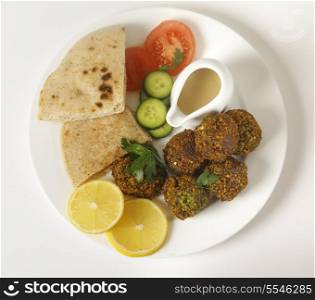 Homemade falafels (herbed and spicy chickpea balls) on a plate with Egyptian flat bread, lemon slices, tomato, cucumber and a tahina sauce, viewed from above