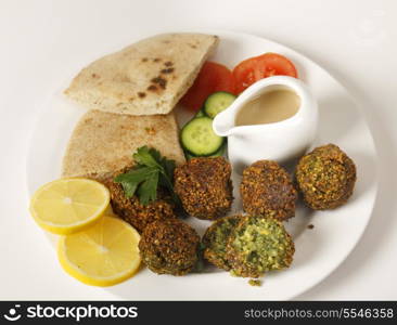 Homemade falafels (herbed and spicy chickpea balls) on a plate with Egyptian flat bread, lemon slices, tomato, cucumber and a tahina sauce.