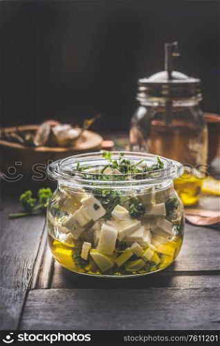 Homemade feta cheese cubes in jar with olives oil and herbs on dark rustic table background. Marinated feta cheese. Tasty food. Country style.