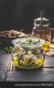 Homemade feta cheese cubes in jar with olives oil and herbs on dark rustic table background. Marinated feta cheese. Tasty food. Country style.