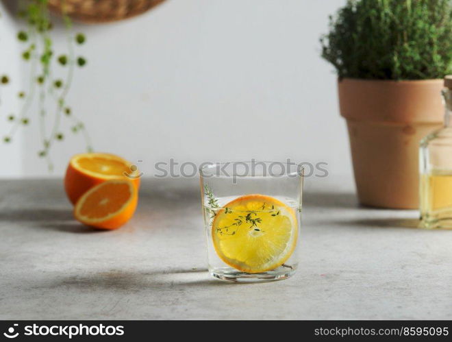Homemade flavored water with orange slice and thyme in glass at grey concrete table with fruits and herbs. Refreshing summer drink with citrus fruit. Front view.