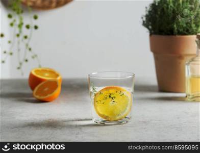 Homemade flavored water with orange slice and thyme in glass at grey concrete table with fruits and herbs. Refreshing summer drink with citrus fruit. Front view.
