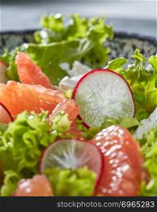 Homemade freshly salad from natural organic vegetables, fruits, cheese. Concept of healthy diet food. Close-up view with soft focus.. Close-up of fresh natural ingredients of vegetarian salad - round slices of radish, pieces of grapefruit, green lettuce in a ceramic bowl.
