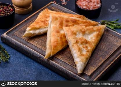 Homemade fried chebureks with meat and herbs on a cutting board on a dark concrete background, traditional Caucasian cuisine. Homemade fried chebureks with meat and herbs on a cutting board