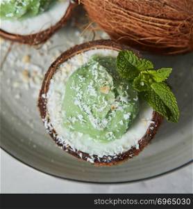 Homemade green ice cream in a coconut shell, mint leaf on a gray ceramic plate on a light gray. Top view. Vegetarian concept of diet eating. Close-up view a half of coconut with green mint ice cream with sprig of mint and coconut chips.