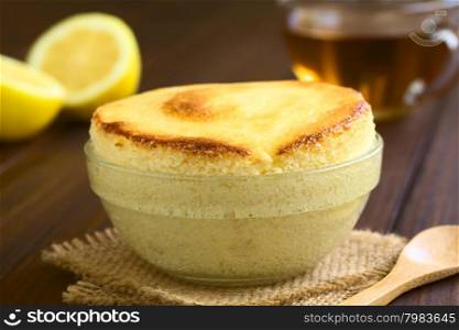Homemade lemon souffle in glass bowl, with cup of tea and lemon halves in the back, photographed on dark wood with natural light (Selective Focus, Focus on the front of the souffle)
