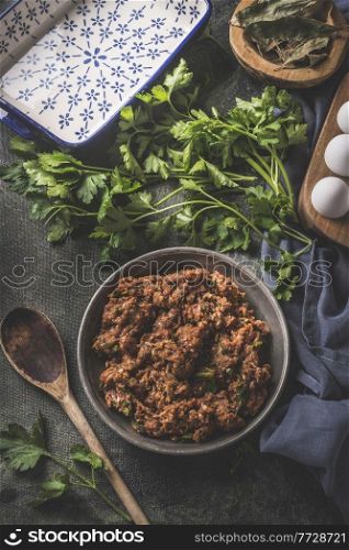Homemade meat stuffing in grey bowl with fresh parsley, herbs, eggs, traditional baking dish and wooden spoon on dark kitchen table. Top view.