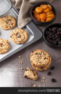 Homemade organic oatmeal cookies with raisins and apricots with baking tray on grey wood background. Black bowl of raisins and dried apricots