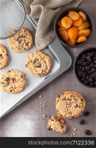 Homemade organic oatmeal cookies with raisins and apricots with baking tray on grey wood background. Black bowl of raisins and dried apricots