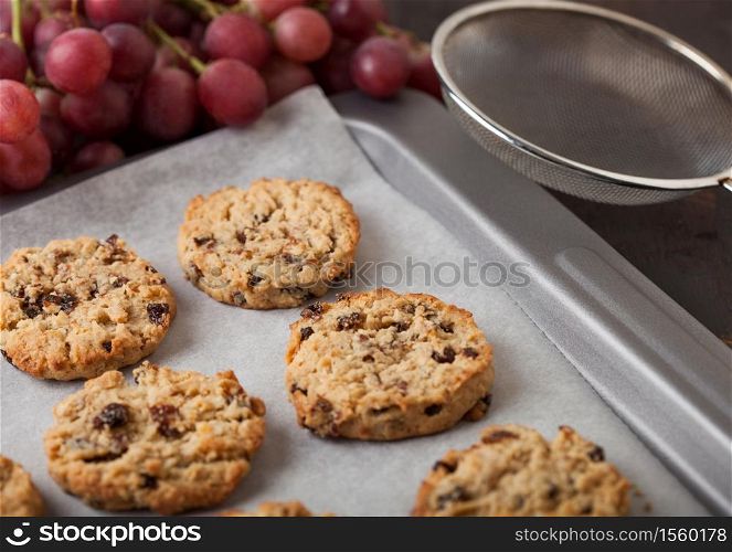 Homemade organic oatmeal cookies with raisins in baking tray with grapes and backing mesh on wooden background.
