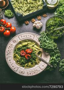 Homemade pasta with raw kale pesto and grilled tomatoes in plate with fork on kitchen table with ingredients. Vegan food. Healthy meal.  Detox vegetables .  Clean eating and dieting concept. Vertical