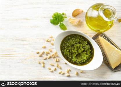 Homemade pesto sauce with ingredients. Sauce pesto in white bowl with basil, olive oil, pine nuts and parmesan cheese on white wooden background. Top view, flat lay with copy space for text.. Homemade pesto sauce with food ingredients on white wooden background