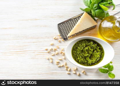 Homemade pesto sause with ingredients. Sause pesto in white bowl with basil, olive oil, pine nuts and parmesan cheese on white wooden background. Top view, flat lay with copy space for text.. Homemade pesto sause with food ingredients on white wooden background