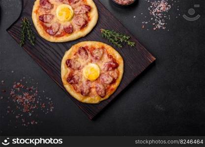 Homemade pizza with sausages, tomatoes, cheese, spices and herbs on a wooden cutting board against a dark concrete background. Homemade pizza with sausages, tomatoes, cheese, spices and herbs on a wooden cutting board