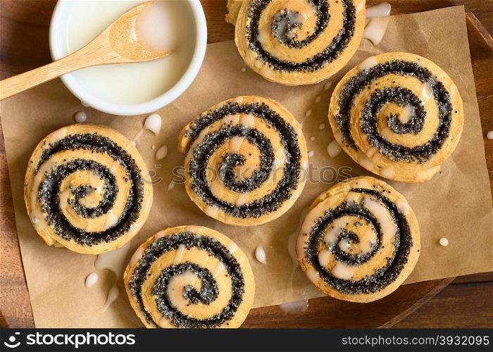 Homemade poppy seed rolls with icing on wooden plate, photographed overhead with natural light (Selective Focus, Focus on the top of the rolls)