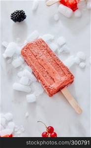 Homemade popsicle blueberry and cream in color of the year 2019 Living Coral Pantone on crushed ice over a marble background with berries, top view. Summer concept. Berry black currant ice cream sorbet popsicles in color of the year 2019 Living Coral Pantone on crushed ice over a marble background with berries, top view