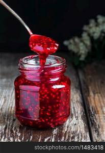 Homemade raspberry jam in a glass jar on a wooden table. Close-up, selective focus. Spoon with jam over the jar, jam dripping from a spoon. Homemade raspberry jam in a glass jar on a wooden table. Close-up, selective focus.
