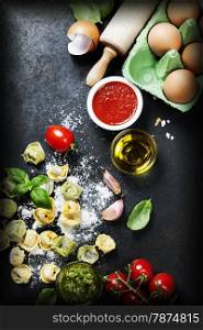 Homemade raw Italian tortellini, basil leaves with flour and tomatoes on dark vintage background