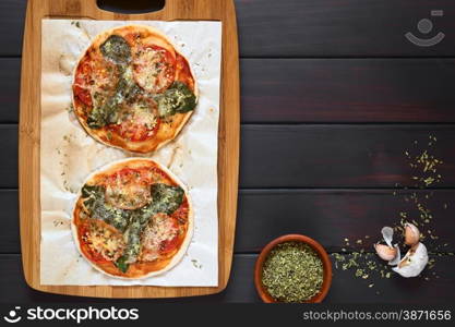 Homemade spinach and tomato pizza on baking paper on wooden board, dried oregano and garlic on the side, photographed overhead on dark wood with natural light