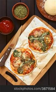 Homemade spinach and tomato pizza on baking paper on wooden board, pizza dough, dried oregano, tomato sauce on the side, photographed overhead on dark wood with natural light
