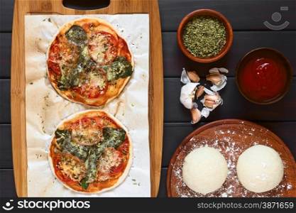 Homemade spinach and tomato pizza on baking paper on wooden board, pizza dough, dried oregano, tomato sauce, garlic on the side, photographed overhead on dark wood with natural light